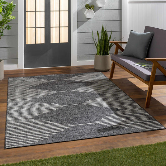Gray patterned rug on a wooden deck with a chair and plants in the background.