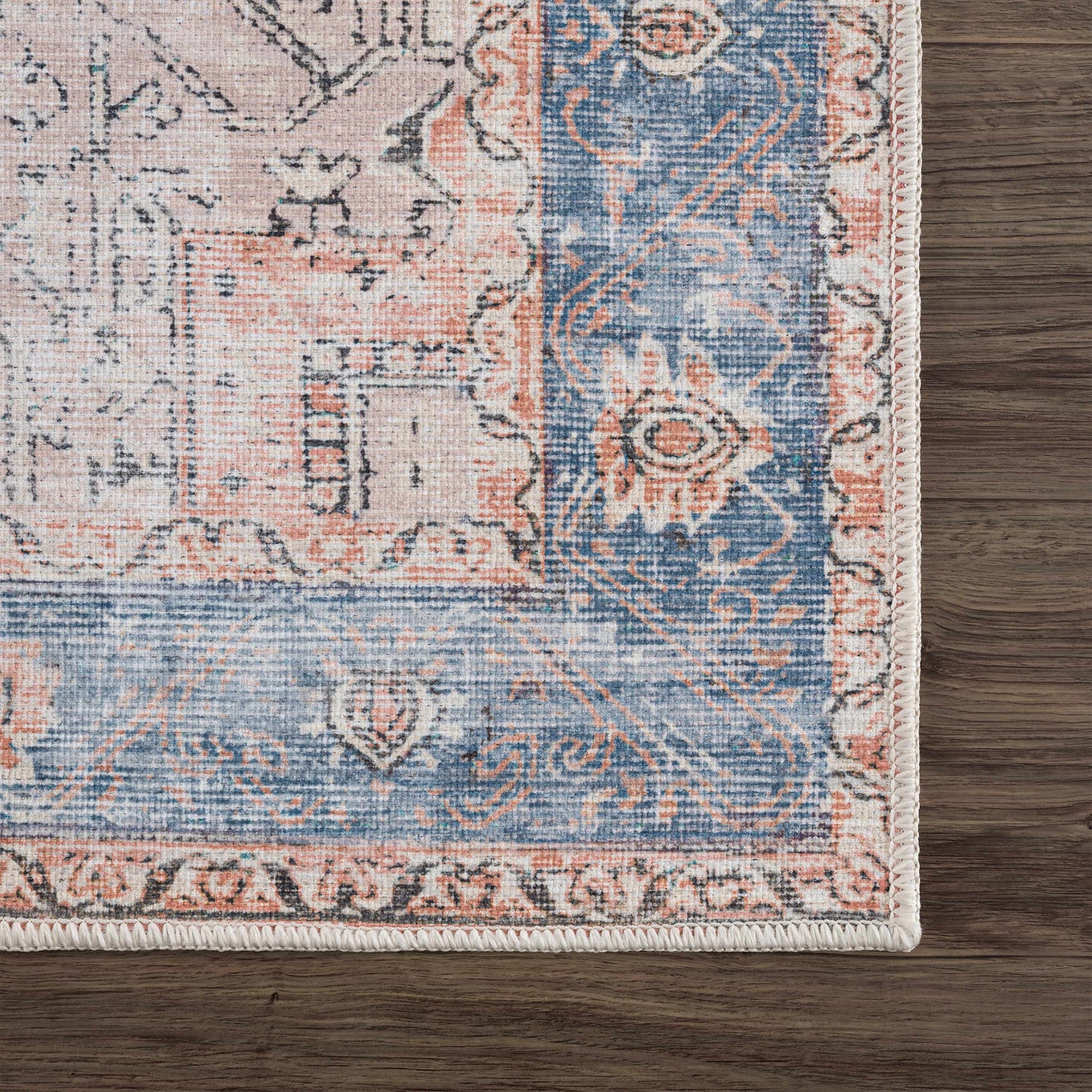Corner of a patterned rug with blue, pink, and beige colors on a wooden floor.