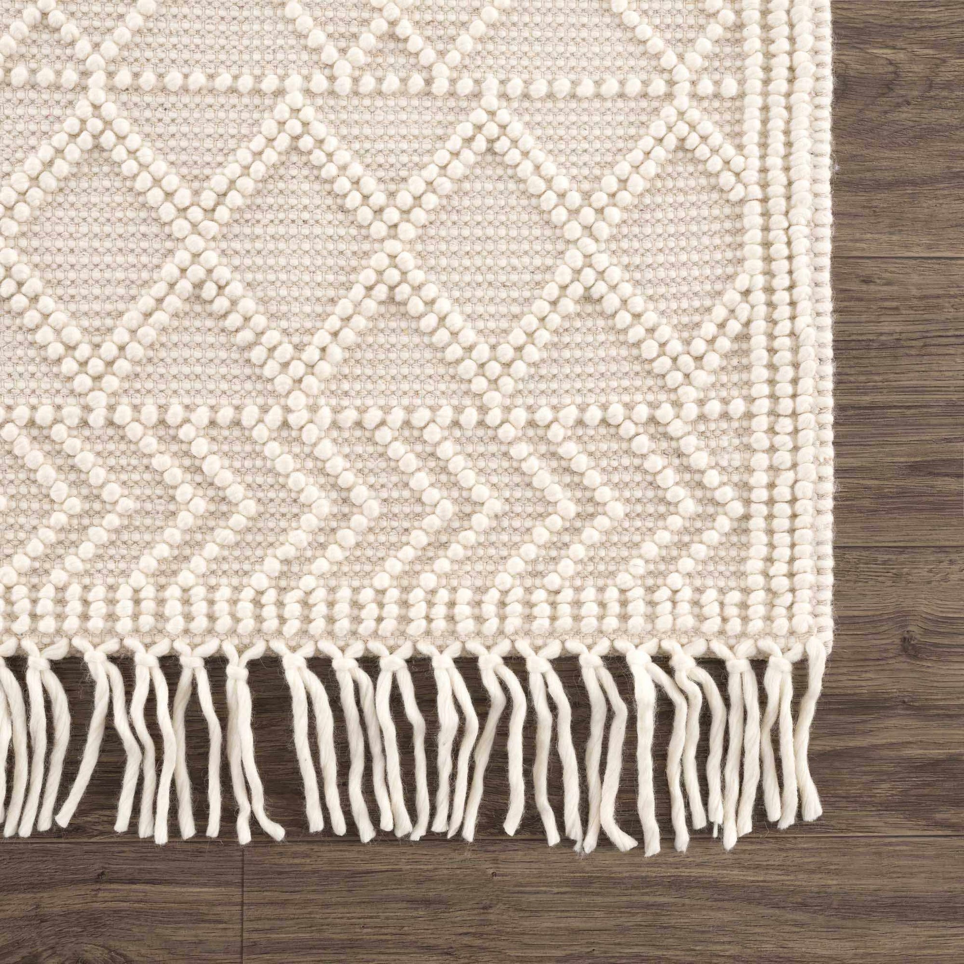 Beige woven rug with diamond pattern and white fringe on a wooden floor.