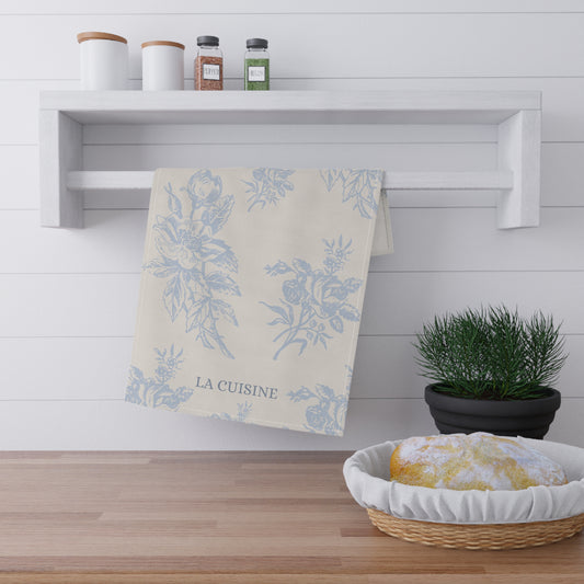 Kitchen towel with floral design hanging on a shelf above a wooden counter with a basket of bread.