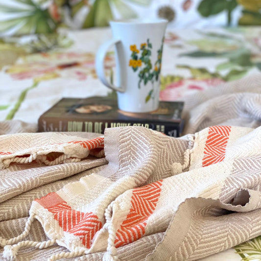 Beige and rust throw blanket styled with book and floral mug on printed bedding