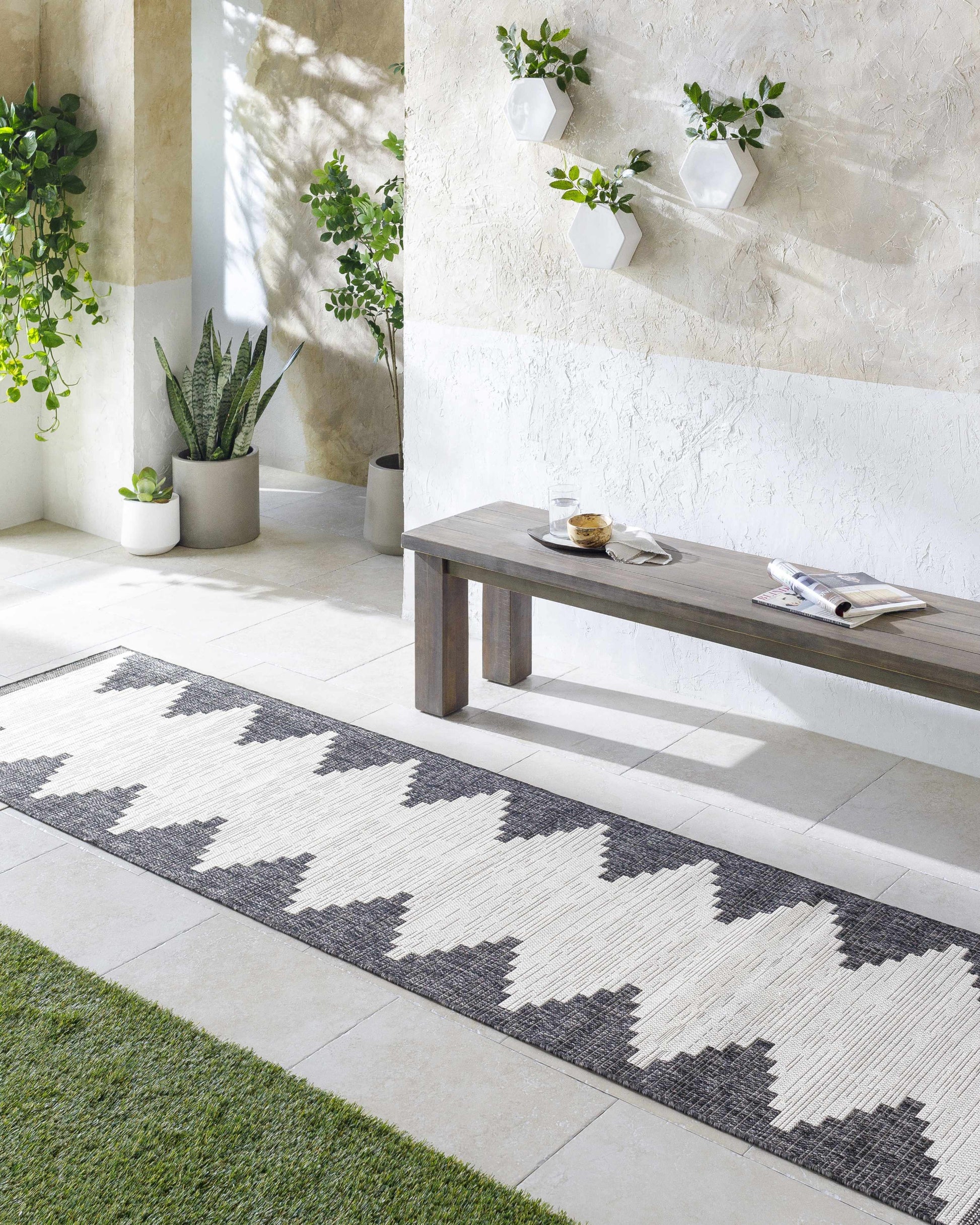 Modern outdoor patio with a geometric patterned rug, wooden bench, and potted plants.
